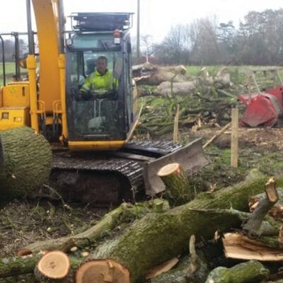 A yellow excavator being operated by a Highfield Tree Care professional to move and clear large felled tree trunks on a muddy development site at Oldham.