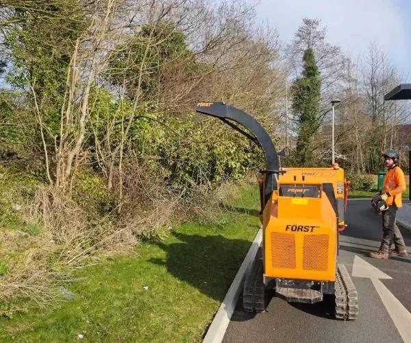A Highfield tree surgeon in high-visibility orange safety gear and a helmet standing next to a yellow Forst tracked wood chipper on a paved path in Lancashire while clearing storm-damaged trees and branches.